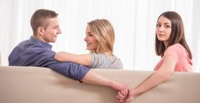 Love triangle. Beautiful young woman holding hands with man sitting near his girlfriend on sofa.